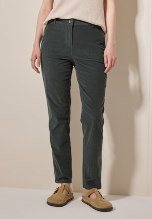 Femme portant un pantalon slim vert foncé et des chaussures beige en daim à enfiler, debout contre un mur clair.