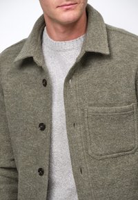 Man som bär en strukturerad olivgrön knäppt ulljacka över en ljusgrå stickad tröja, som visar övre delen av bröstet och kragens område.