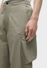 Lettere, olivenfarvede cargo-bukser med en tekstureret finish, der har en sidelomme og en knaplukning. Simpelt design og funktionelt detalje.