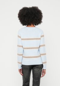 Moteris su garbanotais plaukais vilki šviesiai mėlyną megztinį su smėlio spalvos horizontaliomis juostelėmis ir juodas tekstūruotas kelnes, žiūrintį tolyn nuo kameros.