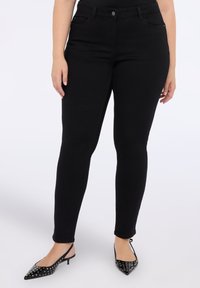 Mujer que lleva unos pantalones skinny de cintura alta negros y unos tacones de slingback puntiagudos negros con tachuelas, de pie frente a un fondo blanco.