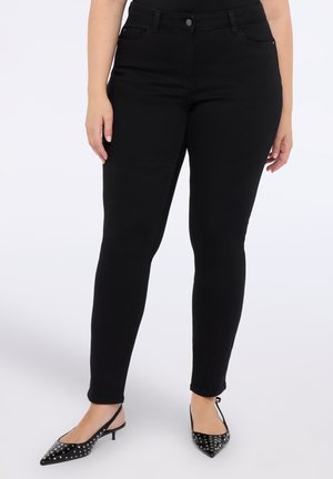 Femme portant un jean skinny taille haute noir et des escarpins à bout pointu noirs à talon slingback cloutés, se tenant contre un fond blanc.