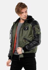 Olivgrüne Bomberjacke mit schwarzen Lederärmel, ausgestattet mit einem orangefarbenen Innenfutter, mehreren mit Reißverschlüssen versehenen Taschen und einem Aufnäher auf der Brust.