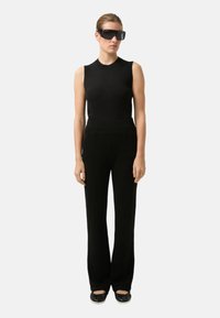 Schwarzes ärmelloses Oberteil, hoch taillierte schwarze Hose mit Taschenähnlichen Nähten und schwarze Slip-On-Schuhe. Modell trägt übergroße schwarze Sonnenbrillen.