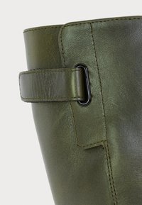 Gros plan sur la tige d'une botte en cuir vert olive, présentant une sangle cousue passant à travers une boucle ovale en métal noir.