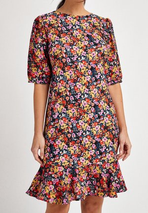 Femme portant une robe courte aux genoux, manches courtes, avec imprimé floral multicolore et ourlet à volants, sur fond uni.