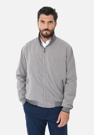 Uomo con capelli scuri e barba che indossa una giacca grigia chiaro con cerniera sopra una maglietta bianca e pantaloni blu scuro, in piedi contro uno sfondo bianco.