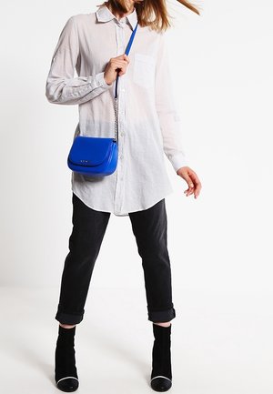 White button-up shirt with long sleeves, paired with black rolled-up jeans and black ankle boots. A small blue crossbody bag hangs.