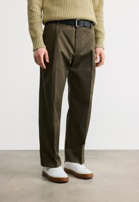 Άνδρας που φοράει ελαιστι πράσινο παντελόνι ίσιας γραμμής, μαύρη ζώνη, μπεζ πλεκτό πουλόβερ και λευκά αθλητικά παπούτσια με σόλες από καουτσούκ, στέκεται σε γκρι πάτωμα.