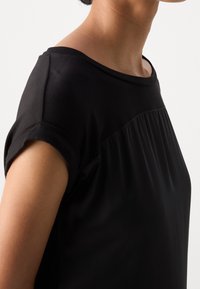 Mustapaita, jossa on lyhyet hihat, pyöreä pääntie ja poimutettu yksityiskohta edessä. Suunniteltu kevyestä, sileästä kankaasta ja rento istuvuus.