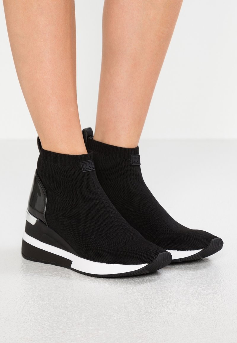 Jambes portant des baskets compensées en maille noire avec des accents blancs et un panneau de talon noir brillant sur un fond de sol clair.