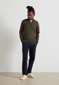 Olijfgroen pullover met een half-rits ontwerp, geribbelde textuur en klein geborduurd logo; gecombineerd met marineblauwe broek met platte voorkant en crèmekleurige sneakers.