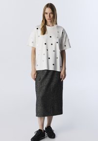 Weißes Kurzarmoberteil mit metallischen Punkten, kombiniert mit einem strukturierten dunkelgrauen Midirock und schwarzen Turnschuhen, was ein kontrastierendes Outfit ergibt.