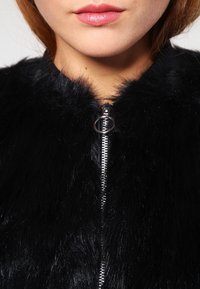 Svart fuskpälsjacka med hög krage, med en silverdragkedja framtill och en mjuk, texturerad yta.
