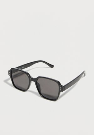 Lunettes de soleil rectangulaires noires avec des verres foncés et des branches épaisses reposant sur une surface blanche.