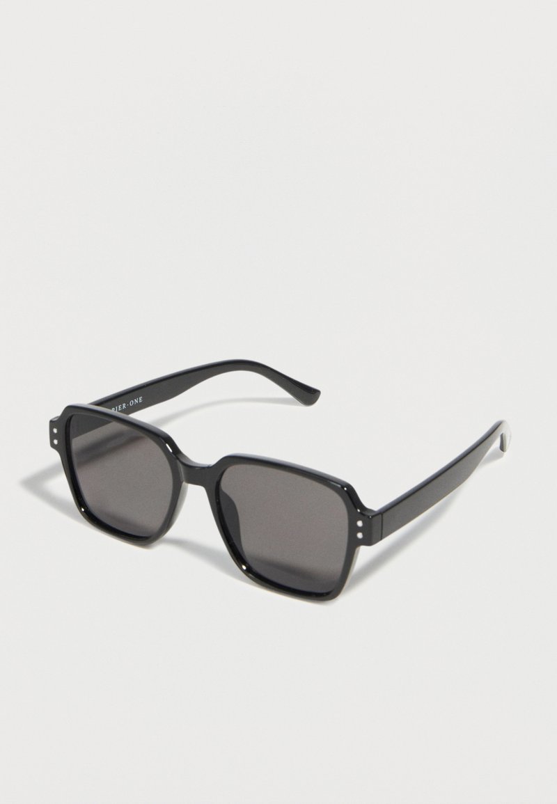 Μαύρα ορθογώνια γυαλιά ηλίου με σκούρους φακούς και χοντρές βάσεις που ακουμπούν σε λευκή επιφάνεια.