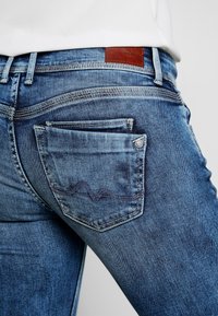 Modré denimové džíny s středním pasem, s detailním prošíváním zadních kapes a hnědou koženou nášivkou v blízkosti pasu.