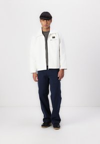 White jacket with a collar and zip closure, worn over a gray shirt, paired with navy pants and black sneakers. Simple design.