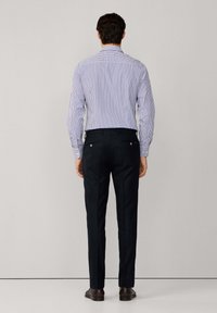 Prugasta plava i bijela košulja dugih rukava s kragnom na gumbe, uparena s tamnim hlačama. Glatka tkanina, prilagođeni kroj i vidljiv detalj na leđima.