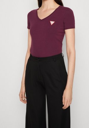 Femme portant un t-shirt violet près du corps à col en V avec un petit logo triangle rouge et blanc et un pantalon noir taille haute.