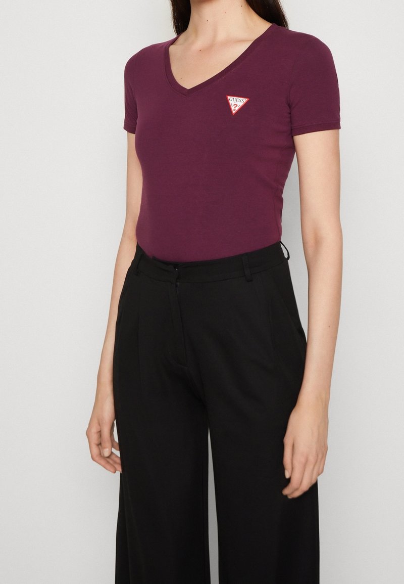 Femme portant un t-shirt violet près du corps à col en V avec un petit logo triangle rouge et blanc et un pantalon noir taille haute.