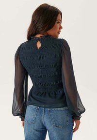 Námořnická modrá blůza s nařaseným výstřihem, průhlednými balónovými rukávy a detailem v podobě klíčové dírky vzadu, spárovaná s modrými džíny. Hladký povrch a strukturovaný střih.