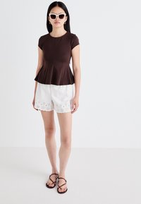 Brown peplum top paired with white shorts featuring a scalloped hem and eyelet detailing. Model wears sunglasses and flat sandals.