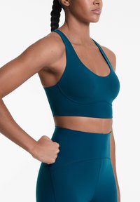 Soutien-gorge de sport teal avec un col en scoop, des bretelles larges et une large bande sous la poitrine. Matériau lisse et extensible conçu pour le confort.