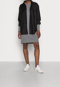 Camicia nera a bottoni sopra un vestito grigio, entrambi realizzati in tessuto morbido. La camicia ha due tasche frontali e maniche lunghe. Sneakers bianche completano il look.