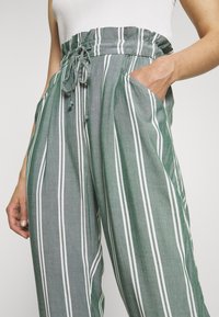 Pantalon léger à rayures vertes avec taille élastique, cordon de serrage et poches latérales. Rayures blanches verticales et texture douce.