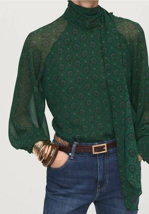 Woman wearing sheer dark green patterned blouse with high neck and large bow, blue jeans, brown belt, and stacked gold bangles on wrist.