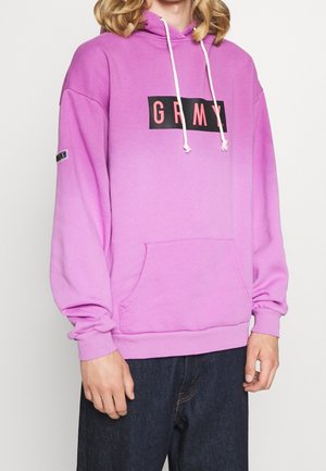 Osoba ubrana w luźną różową bluzę z kapturem z przednią kieszenią, białymi troczkami oraz czarnym prostokątnym logo z czerwonymi literami "GRMY", w połączeniu z ciemnymi dżinsami.