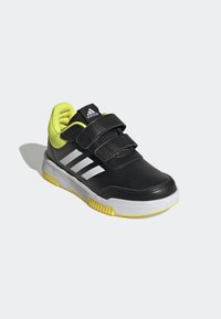 Zapatilla deportiva negra con dos correas ajustables de velcro, tres franjas blancas y un detalle amarillo en el talón y el cuello. Suela exterior de goma.