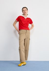 Rotes Sportshirt mit weißem Kragen und gelben Akzenten, kombiniert mit beigen Cargo-Hosen und gelben Sportschuhen. Vor einem schlichten Hintergrund.
