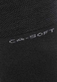 Zwarte sokken van zacht materiaal met een gebreide textuur. Heeft grijze bedrukte tekst langs de elastische rand die "AS F. 90/10" zegt.