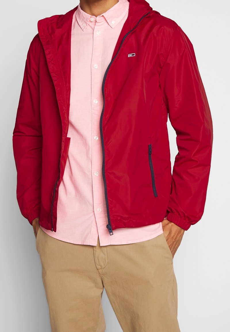 Veste en nylon rouge avec capuche, poignets élastiques et poche zippée, portée par-dessus une chemise à boutons rose clair.