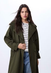Oliven grøn trenchcoat med dobbelte knapper, bred krave og opkrøllede ærmer. Båret over en hvid- og sortstribet skjorte og denimjeans.