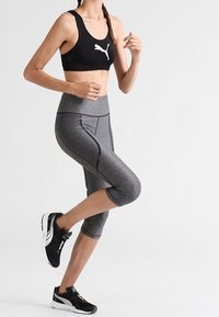 Czarny stanik sportowy z białym logo, szare legginsy z krótkim krojem, z teksturowanym wzorem i szwami, czarne buty sportowe z białymi akcentami.