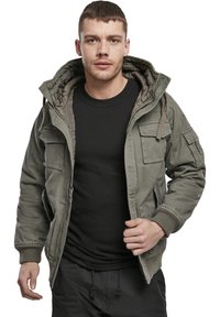 Olivgrüne Bomberjacke mit Kapuze, ausgestattet mit Fronttaschen, elastischen Bündchen und einem glatten Innenfutter. Über einem schwarzen Hemd getragen.
