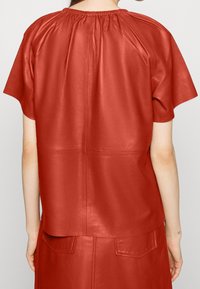 Haut en cuir rouge avec un col froncé et des manches courtes, offrant une texture lisse et une coupe carrée. Détail de couture au dos visible.