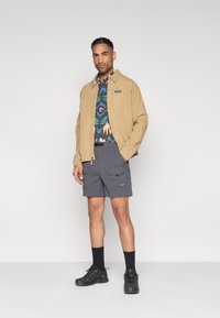 Καφέ σακάκι, πουκάμισο με σχέδια σε πράσινο και μπλε, γκρι σορτς cargo, μαύρες κάλτσες και μαύρα αθλητικά παπούτσια. Το υλικό φαίνεται ελαφρύ με λειτουργικό σχεδιασμό.