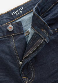 Jeans de denim de corte ajustado en azul oscuro. Presentan un cierre de cremallera, botón de cobre y costuras naranjas en contraste. La etiqueta indica "34 R."
