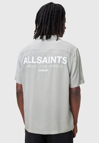 Szara koszulka krótki rękaw o luźnym kroju, z dużym białym napisem "ALLSAINTS LONDON" z tyłu. Gładka tekstura materiału.