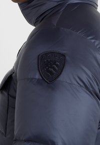 Marinblå puffjacka med hög krage, quiltad textur och en svart broderad badge på vänster axel.
