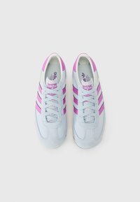 Ljust blå mockaskor med rosa ränder, utrustade med runda snören och en texturerad tåhuvud. Tumdelen visar Adidas-logotypen.