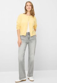 Gelber Cardigan mit Knöpfen, getragen über einem weißen Top. Gepaart mit hellgrauen Jeans und weißen Sneakers. Glatte Textur, lockere Passform.