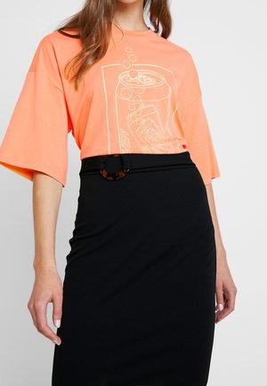 Nainen, jolla on päällään löysä korallinvärinen t-paita, jossa on juomapurkkikuvio, ja korkeavyötäröinen musta hame, jossa kilpikonnakuosinen vyönsolki.