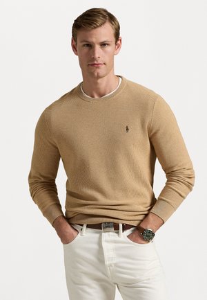 Maglione beige lavorato a maglia con collo e polsini a costine, caratterizzato da un piccolo logo sul petto. Il modello indossa pantaloni bianchi e un orologio da polso, con le mani in tasca.