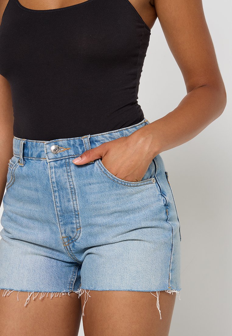 Pantalones cortos de mezclilla en azul claro con dobladillo deshilachado, que cuentan con dos bolsillos laterales y un cierre de botón en la cintura. Textura suave y diseño ajustado.