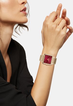 Montre carrée avec un cadran rouge, bracelet en métal doré et design minimaliste. Le bracelet présente des maillons polis et des accents angulaires.